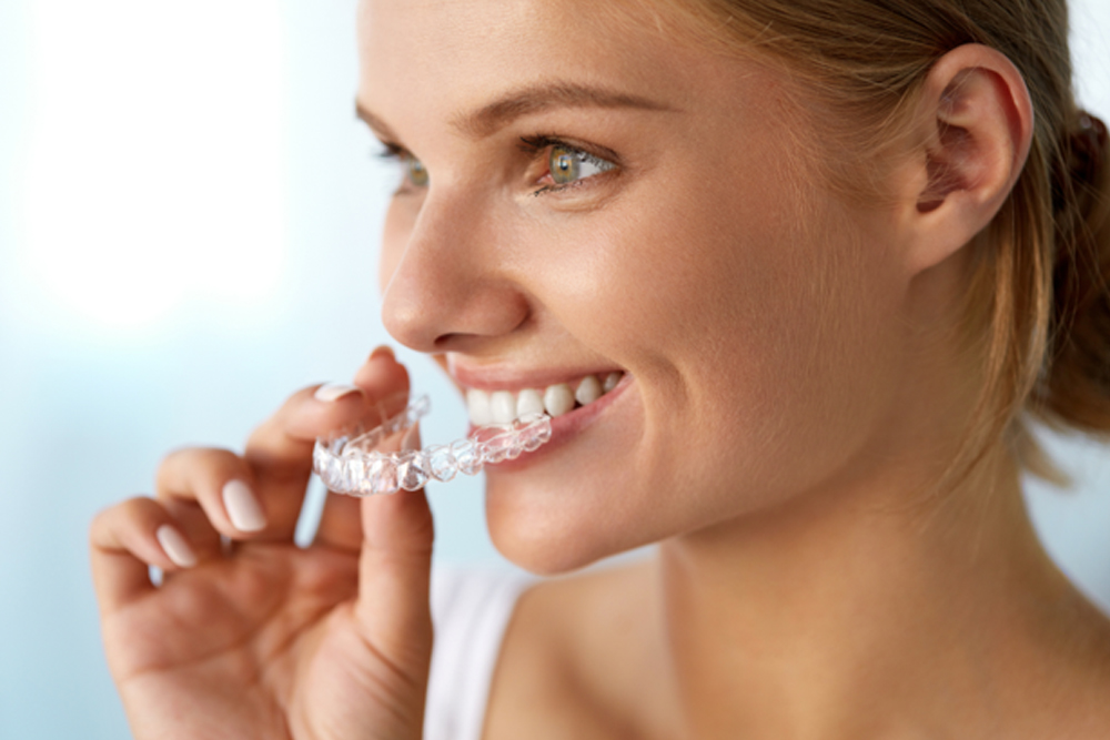 Adult smiling while holding a clear dental aligner near their teeth.